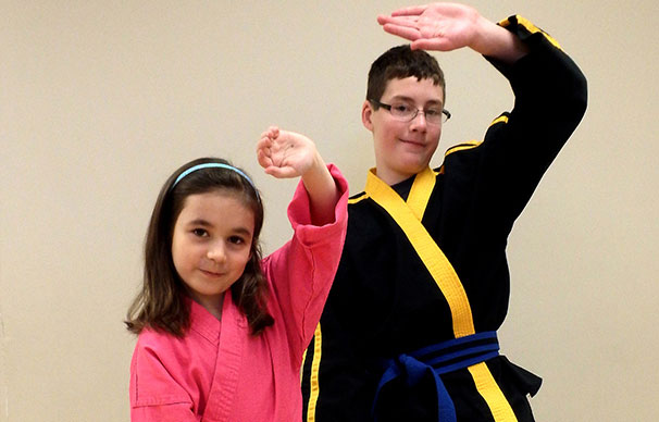 Two siblings are together focused and smiling. The taller, older boy is behind his sister as they do the same stance and pose.